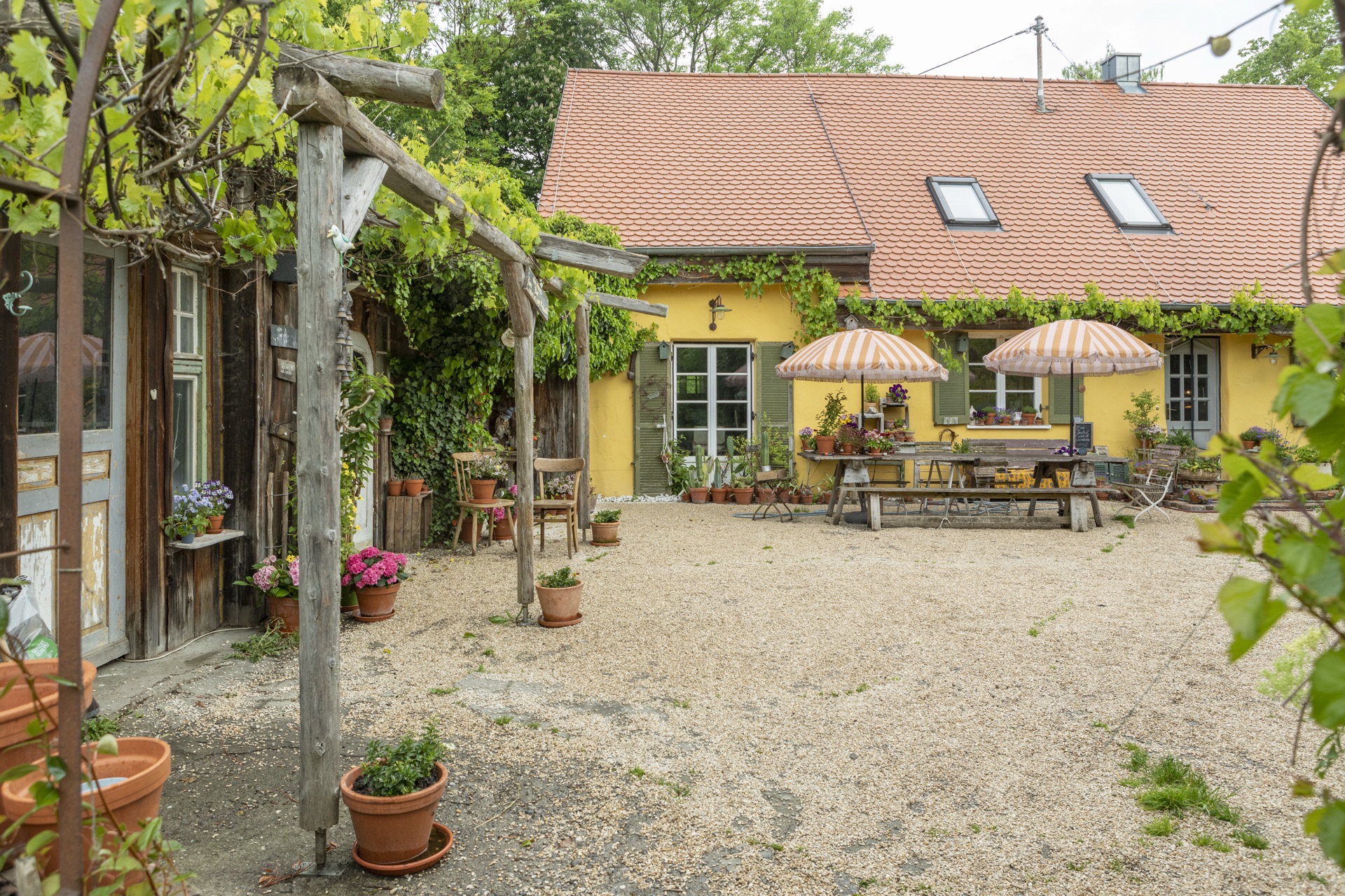 Innenhof des gelben Bauernhauses mit gestreiften Sonnenschirmen, Holztafel und Topfpflanzen unter der Pergola