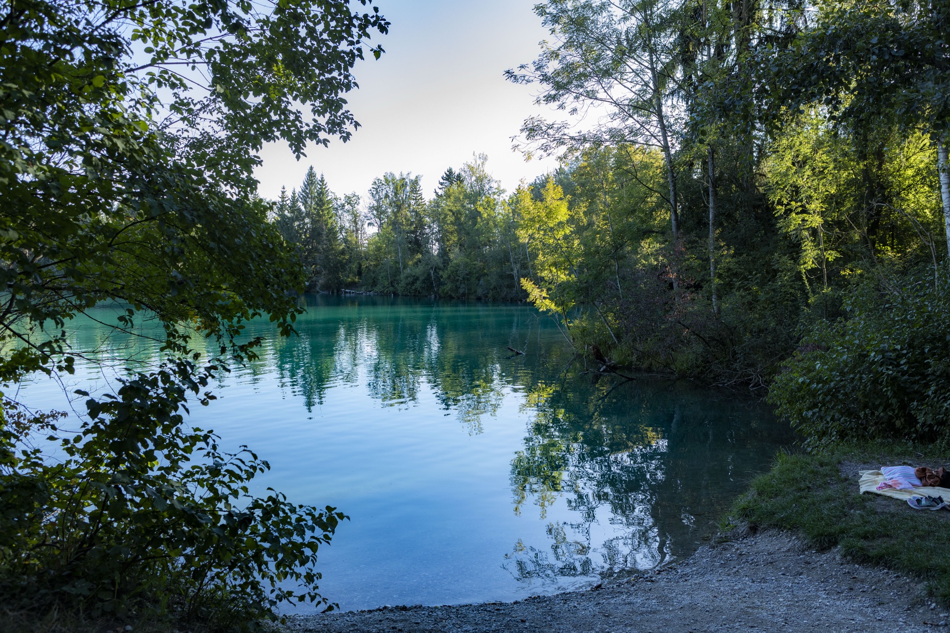Türkisfarbener Baggersee zwischen Bäumen, Handtuch am Kiesufer